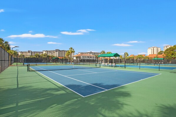 Un court de tennis extérieur doté d’une surface verte et bleue, entouré d’une clôture et d’arbres.