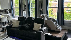 Un salon moderne avec un canapé et des fauteuils massants, de grandes fenêtres avec des rideaux, un coin repas et une cuisine.