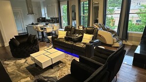 Un salon moderne avec un canapé en cuir noir, une table basse blanche et un fauteuil noir. On aperçoit une cuisine à l’arrière-plan, avec un coin repas.