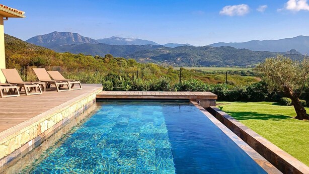 Una piscina cristallina con un pontile in legno e lettini prendisole, immersa nel verde con montagne sullo sfondo.