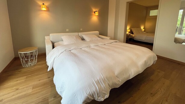 A modern bedroom with a large bed, two wall-mounted lights, a small round side table, and a mirror reflecting the room.