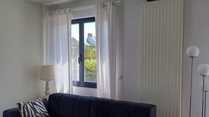 A modern living room with a dark blue sofa, two zebra-patterned pillows, a white table, a floor lamp, and a window with white curtains. - Large, quiet house, large garden, heated swimming pool, ideal for families (La Baule-Escoublac)