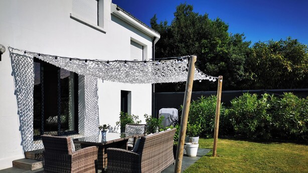 An outdoor patio with wicker furniture, a pergola with string lights, and a white building in the background. - Large, quiet house, large garden, heated swimming pool, ideal for families (La Baule-Escoublac)