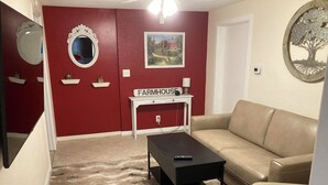 Uma sala de estar com parede vermelha, sofá bege, mesa de centro preta, mesa lateral branca com abajur e uma moldura com quadro.