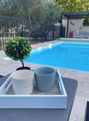 Escena junto a una piscina con una pequeña planta en maceta, dos tazas con texto y una piscina al fondo.