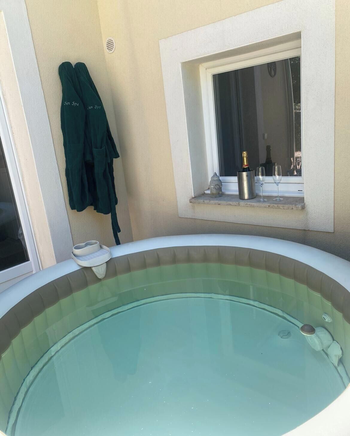 A hot tub with a window above it, a small Buddha statue, and a pair of green towels hanging on the wall.