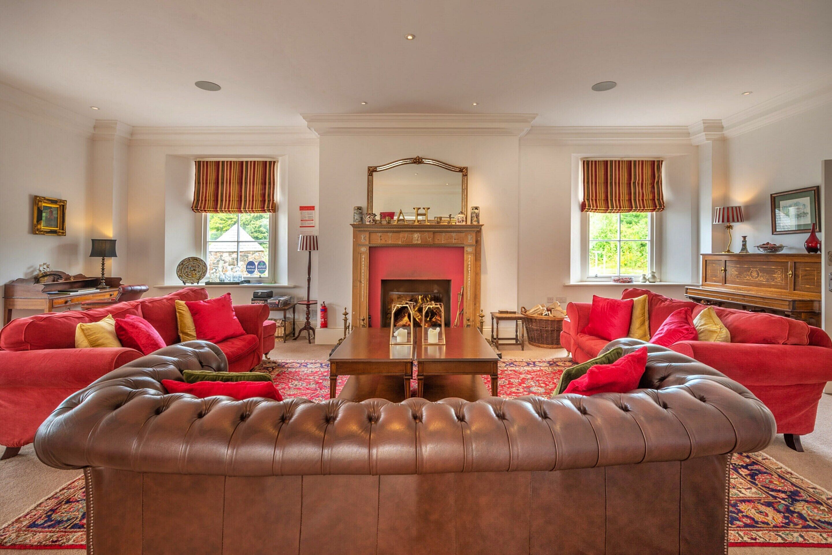 A spacious living room with two red sofas, a brown leather ottoman, a fireplace, and a large mirror above it.