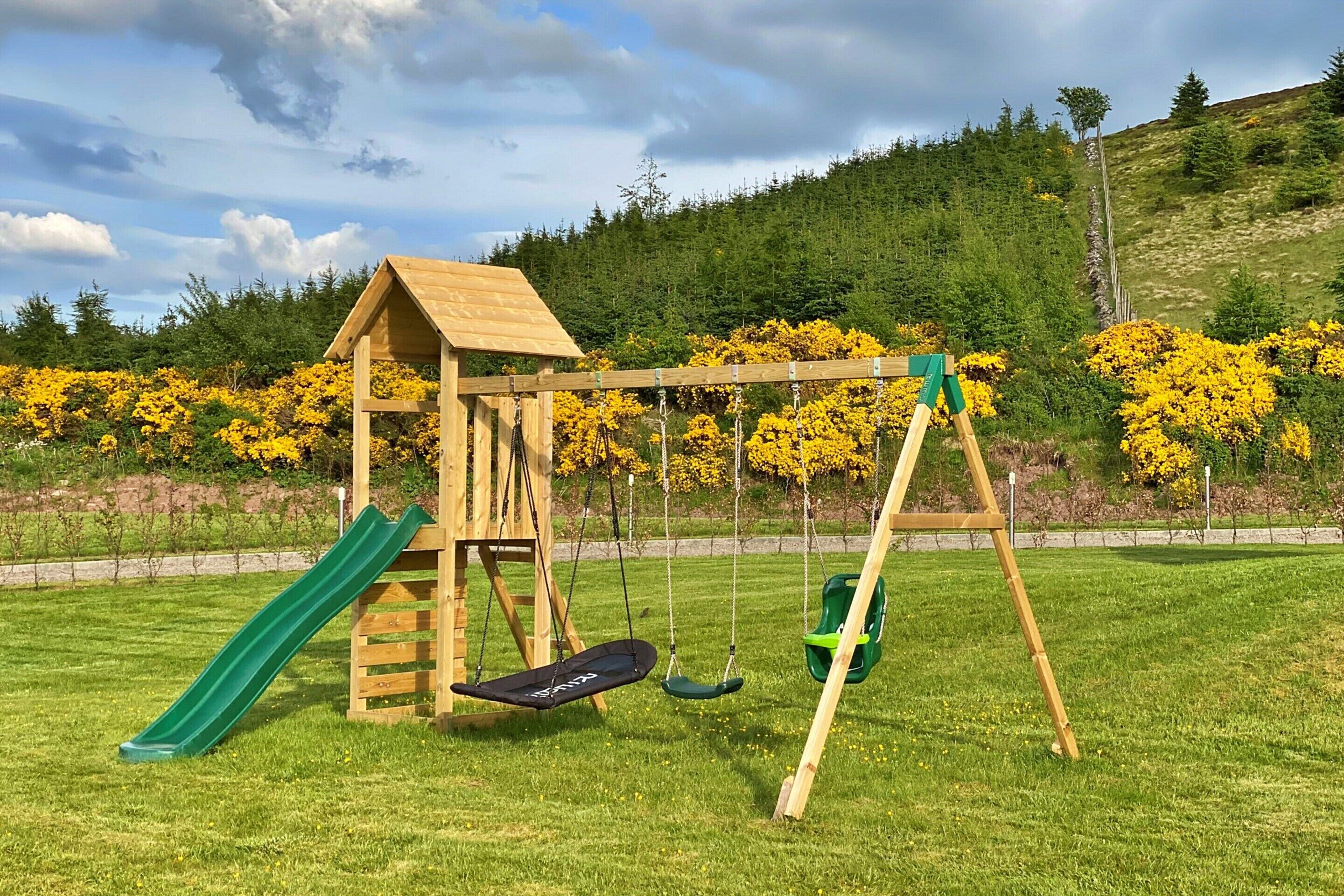 A wooden play structure with a slide and swings in a grassy park area.
