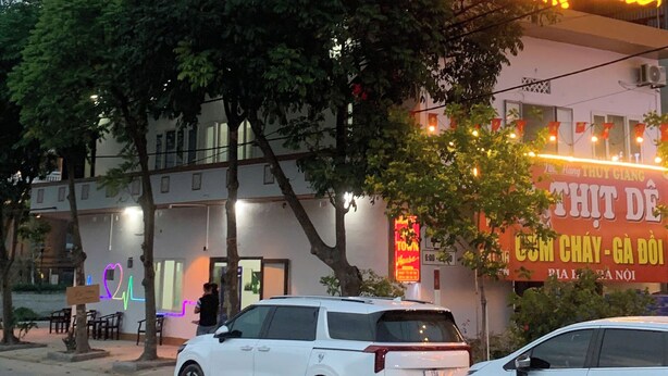 Un edificio con un letrero de neón que dice "HOA LUU GIA TOWN House" y otro letrero que dice "THIT DE". Hay árboles frente al edificio y un coche blanco estacionado en la calle.