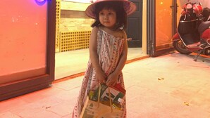 Una niña con un vestido estampado sostiene una bolsa de regalo, parada en un espacio interior con una luz de neón y una patineta en el fondo.