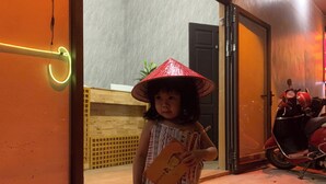 Niño de pie en un umbral, sosteniendo un libro y una mochila, con un patinete estacionado adentro.