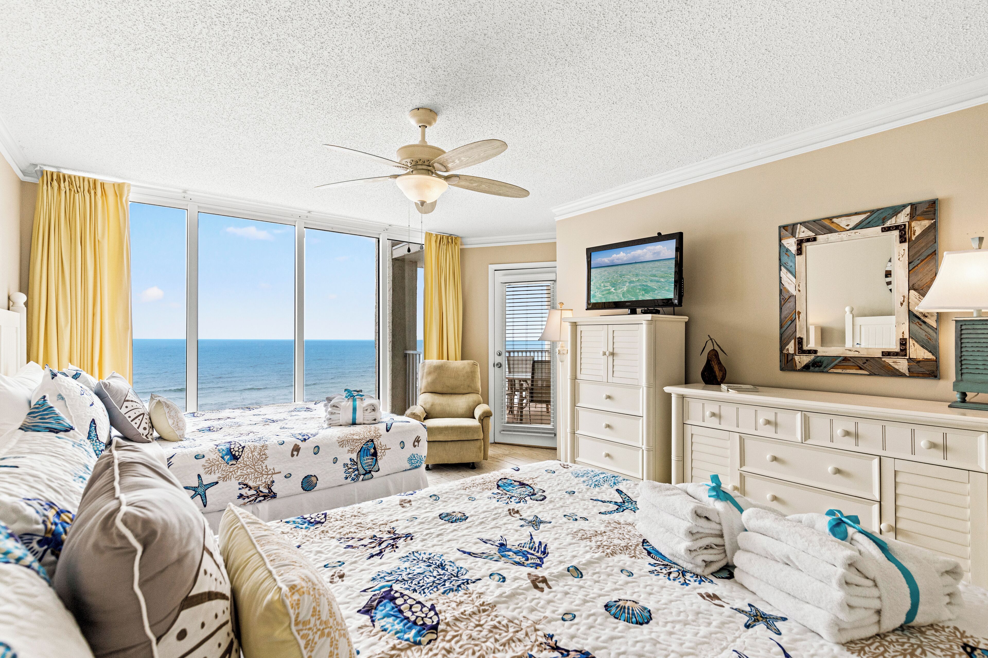 A hotel room with two beds, a TV, a ceiling fan, a large window with a view of the ocean, and a bathroom with a mirror and towel rack.