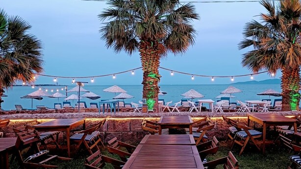 An outdoor seating area with wooden tables and chairs, palm trees, string lights, and umbrellas by the sea. - İMADA MOTEL  (Ayvacik)