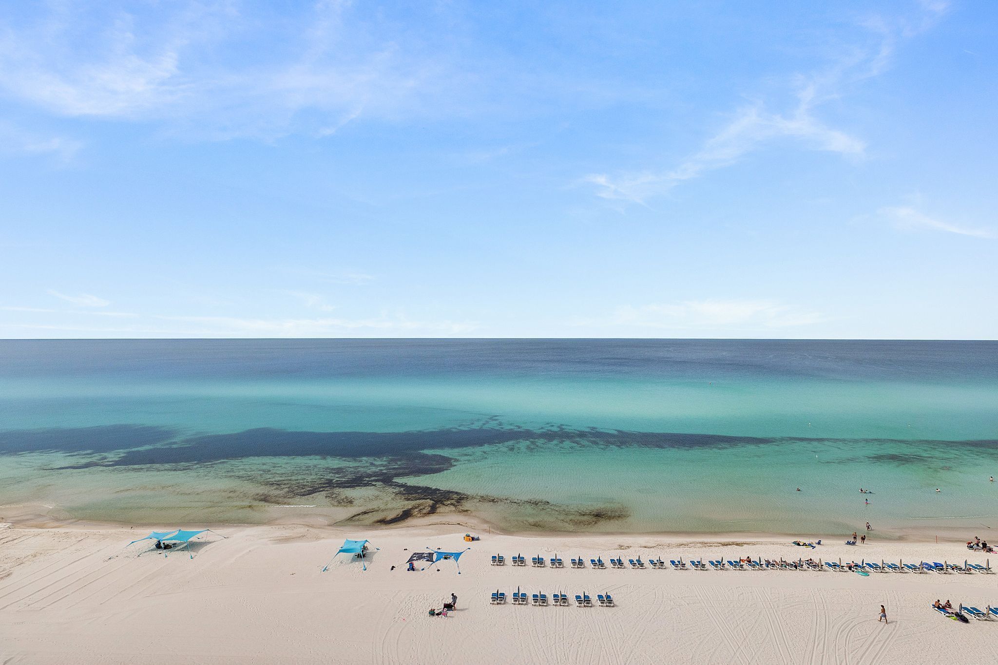 Ein Strand mit Reihen von Liegestühlen und einigen Sonnenschirmen, einem klaren Himmel und ruhiger See.