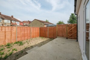 A backyard with a wooden fence, a concrete patio, and a house in the background. - Newly Converted 2BDR 1BA APT in Hounslow (Hounslow)