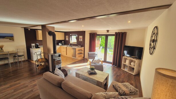 A modern living room with a sofa, coffee table, and TV stand. There is a kitchen area with cabinets and a microwave. The room has a large window with curtains and a view of greenery outside. - Vacation home in Lieper Winkel (Rankwitz)