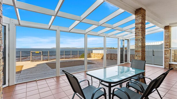 A covered patio with a glass table and chairs, overlooking a beach and ocean. - Sea Breeze @ Barrack Point (Barrack Point)