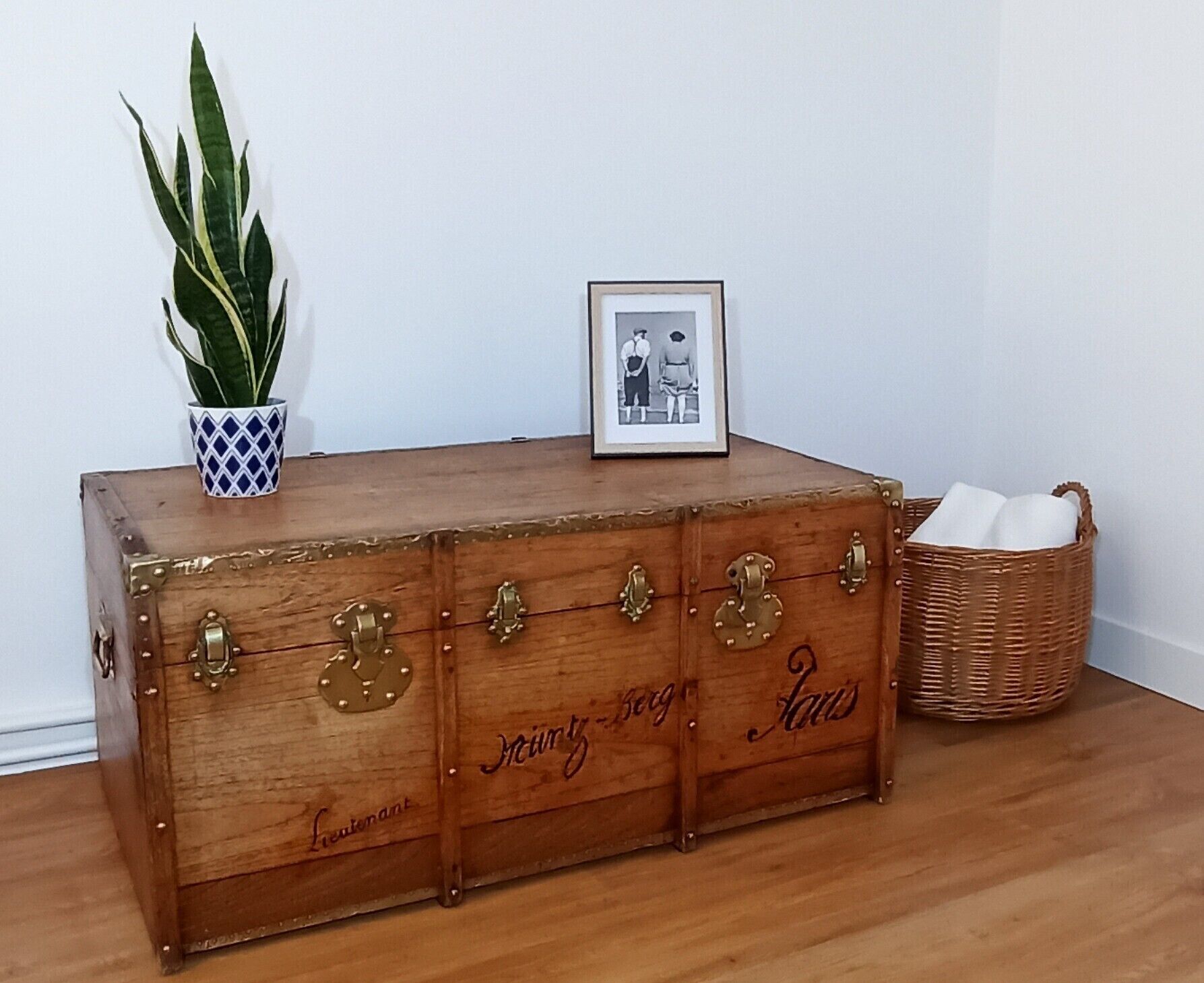 Een houten kist met messing handgrepen en sloten, voorzien van de namen 'Myrtle Berg' en 'Hans', staat op een houten vloer tegen een witte muur.
