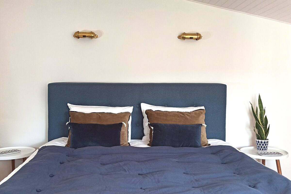 A bedroom with a blue headboard, two bedside tables with lamps, a potted plant, and a bed with a blue duvet and pillows.