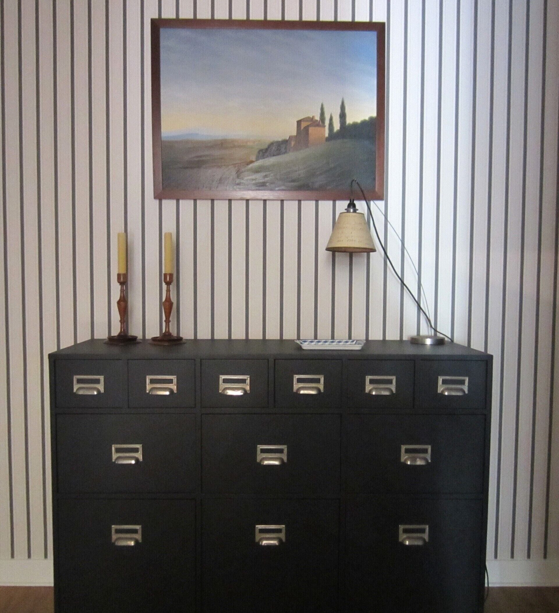 A black filing cabinet with multiple drawers against a striped wall, a framed landscape painting above it, a candle, and a desk lamp.