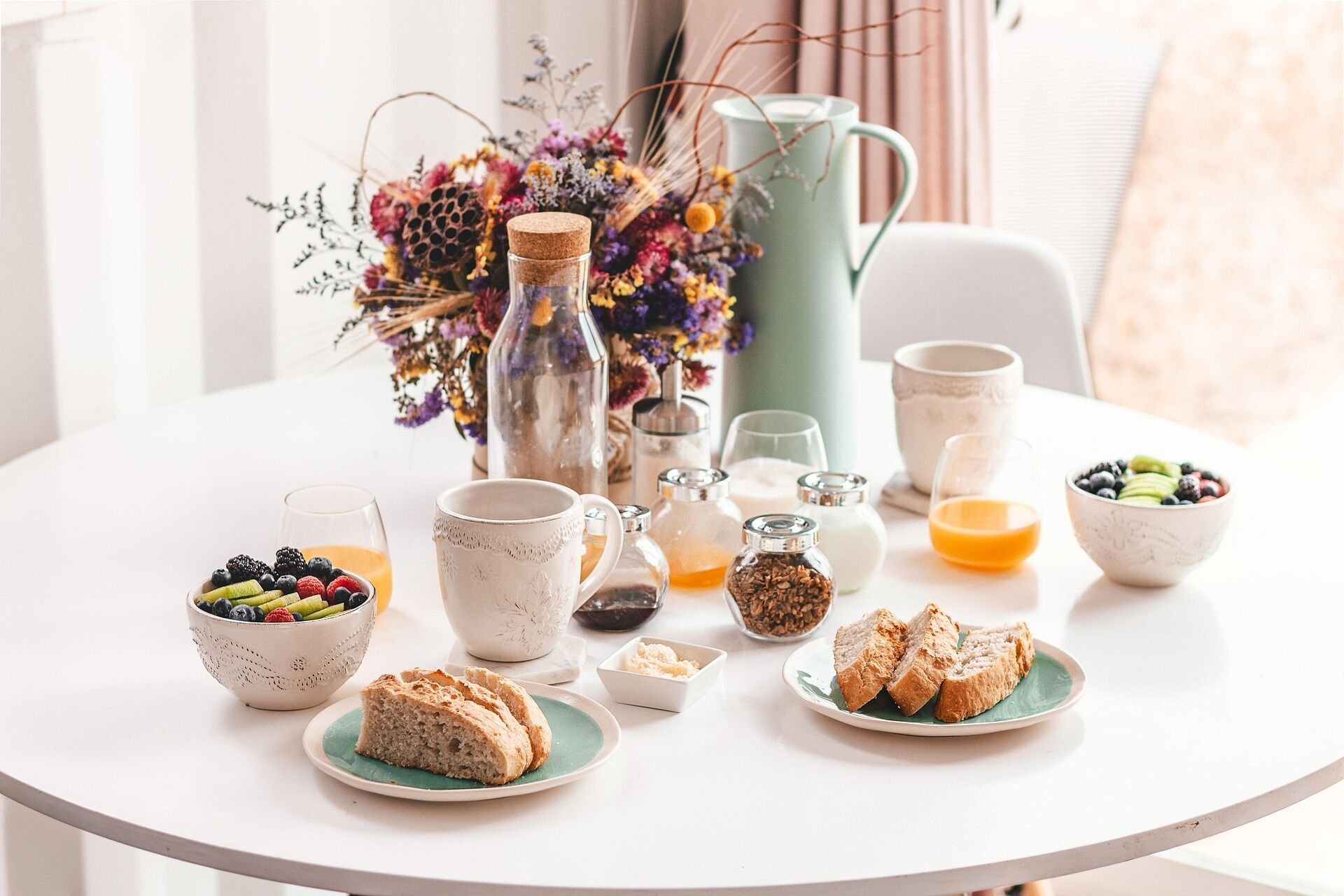 A breakfast table with various food items, including bread, fruits, and beverages, set on a white table in a bright room.