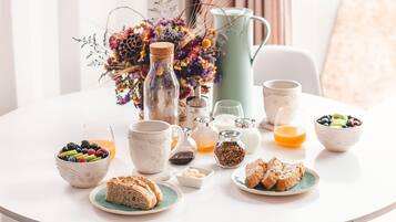 A breakfast table with various food items, including bread, fruits, and beverages, set on a white table in a bright room.