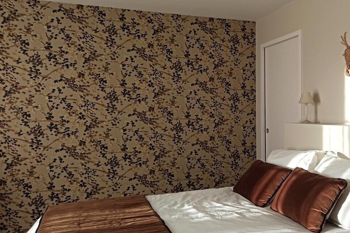 A bedroom with a floral wallpaper, a bed with white and brown bedding, a mounted deer head, and two bedside lamps.