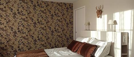 Ein Schlafzimmer mit blumigem Wandpapier, einem Bett mit weißer und brauner Bettwäsche, einem aufgehängten Hirschgeweih und zwei Nachttischlampen.