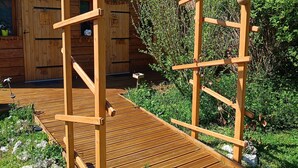 A wooden pergola with a wooden deck, surrounded by greenery and a wooden building in the background. - P'tit ranch SPA PRIVATIF Ô Rêves Atypiques (Boucé)