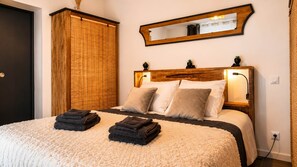 Un lit bien fait, avec deux serviettes pliées, une tête de lit en bois, un miroir fixé au mur et une porte dotée d’un store en bambou.