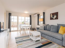 Una sala de estar con un sofá gris, una mesa de centro blanca, un tapete a rayas y un comedor con una mesa y sillas blancas.