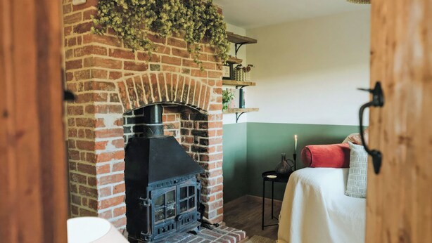A cozy room with a brick fireplace, a bed with a white and red blanket, and a shelf with books and decorative items. - The Green One On The End (Thurton)