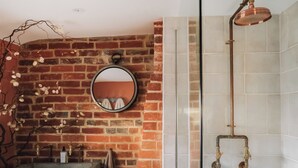 A bathroom with a brick wall, a round mirror, a shower with copper fixtures, and a concrete sink. - The Green One On The End (Thurton)