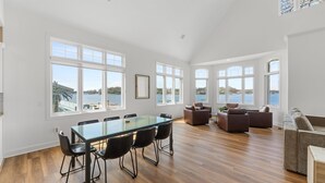 Een ruime woonkamer met een grote glazen tafel en stoelen, een zithoek met sofa’s, en grote ramen met uitzicht op een meer.