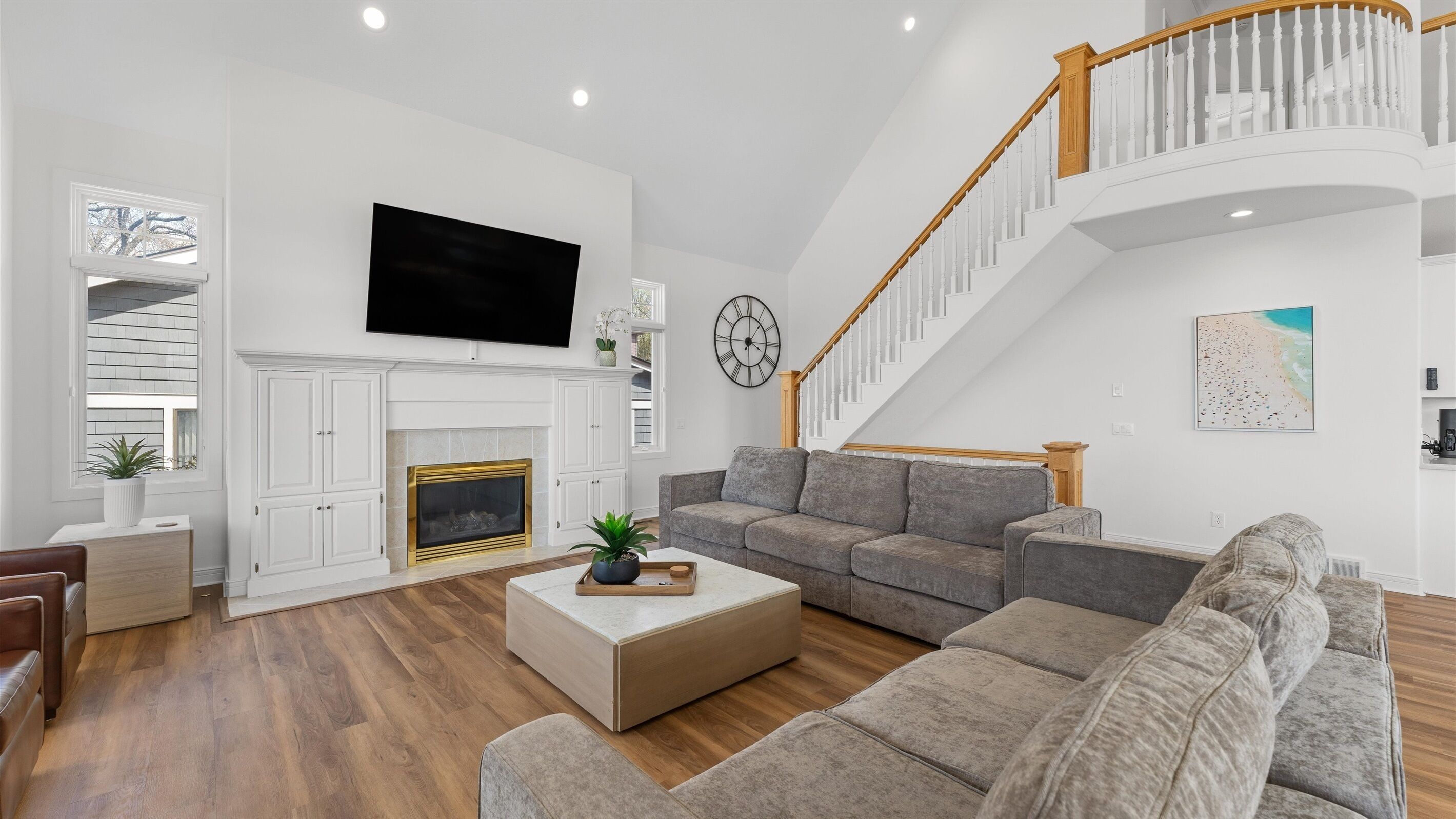 A modern living room with a fireplace, a large flat-screen TV, a staircase, and a beach-themed artwork.