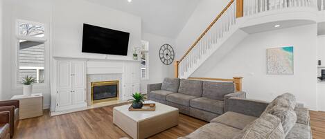 A modern living room with a fireplace, a large flat-screen TV, a staircase, and a beach-themed artwork.