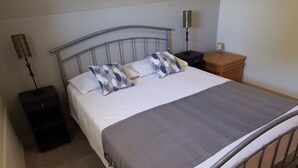 Un lit bien fait, recouvert d’une couverture grise, avec deux oreillers à motifs géométriques, une tête de lit en métal, une table de chevet avec une lampe et une table de nuit en bois.