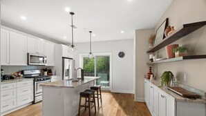 Cocina moderna con muebles blancos, una isla central, electrodomésticos de acero inoxidable y un ventanal con vistas a la vegetación.