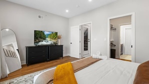 Un dormitorio moderno con cama, televisor en una cómoda, vestidor y baño visible a través de una puerta abierta.