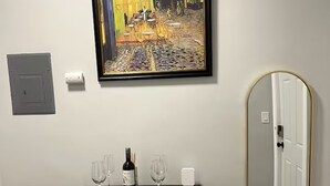 Un aparador negro para vinos con copas y una botella, un cuadro enmarcado encima y un espejo con marco dorado en la pared.
