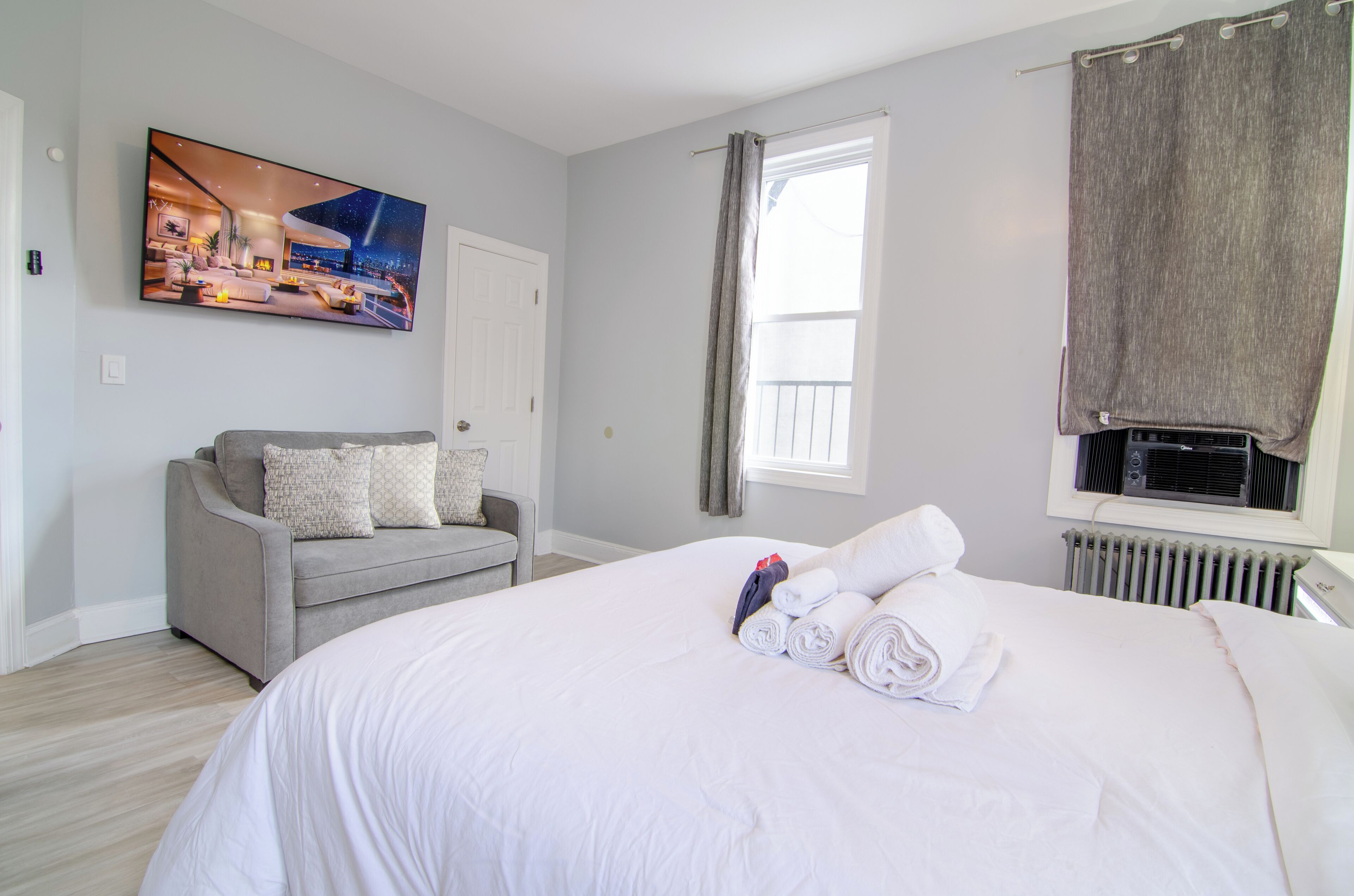 Una habitación de hotel con una cama grande, un sofá gris, un cuadro en la pared, una ventana con cortinas y un radiador.