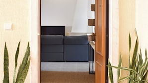 Un couloir mène à une chambre à coucher qui contient un lit, une table de chevet et une lampe. De chaque côté de l’entrée, on trouve une plante en pot.