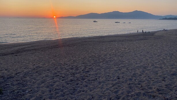 Una playa con un atardecer, mar tranquilo y montañas a lo lejos.