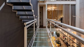 Un intérieur moderne avec un escalier en verre, un balcon au deuxième étage avec un lit, et un coin repas comprenant une table et des chaises.