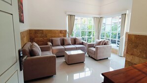 A spacious living room with a beige sofa set, a glass coffee table, and a wooden dining table. Large windows with curtains provide natural light and a view of greenery outside. - 2BR Pool Villa-Twin Flower (Banjar)