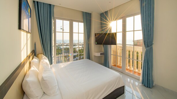 A hotel room with a large bed, a TV mounted on the wall, and a balcony with a view of the sea. - Hoàng Hôn Hotel (Phu Quoc)