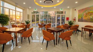 A spacious dining area with multiple tables and orange chairs, large windows with a view of the sunset, and a ceiling with recessed lighting. - Hoàng Hôn Hotel (Phu Quoc)