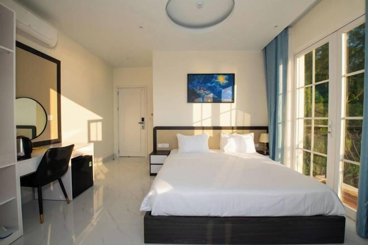Une chambre à coucher moderne avec un grand lit, un bureau avec une chaise, une grande fenêtre et un tableau au mur.