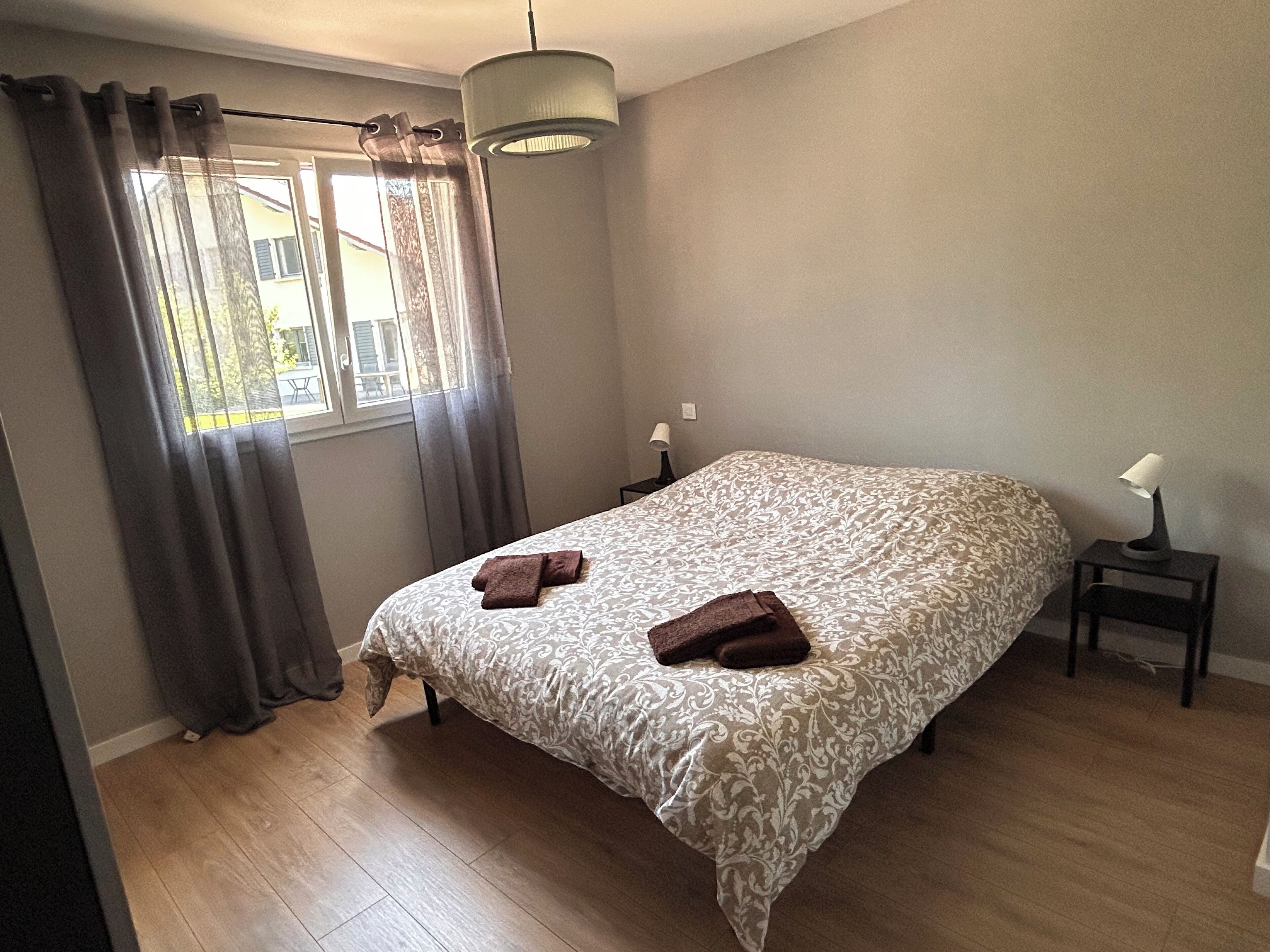 Un dormitorio con cama, mesita de noche, lámpara y ventana con cortinas.