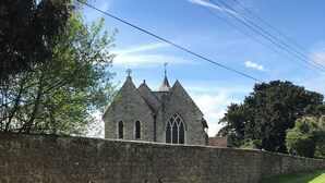 Eine steinerne Kirche mit hohem Spitzhelm und Rundbogenfenstern, umgeben von einer Steinmauer und einer Rasenfläche.